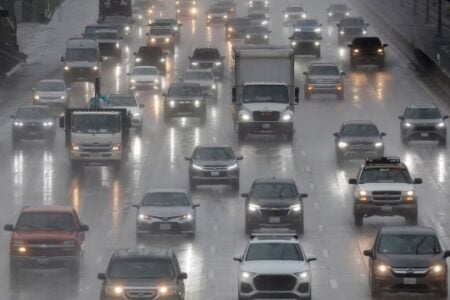 Cars and trucks drive on a wet, multi-lane highway in the rain, with headlights reflecting on the pavement.