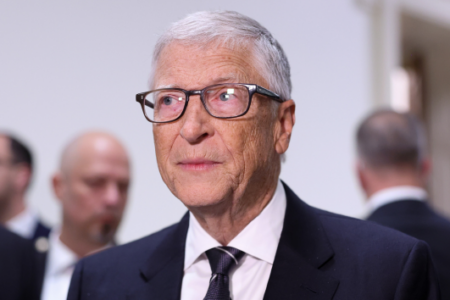 Bill Gates, in a dark suit and glasses, looks toward his left at an indoor event.