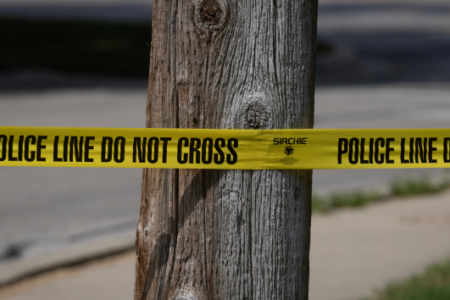 Yellow "POLICE LINE DO NOT CROSS" tape stretched across a weathered wooden pole with a blurred street background.