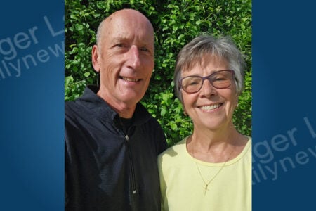 Smiling bald man in black shirt and gray-haired woman with glasses in yellow shirt pose outdoors against green foliage.