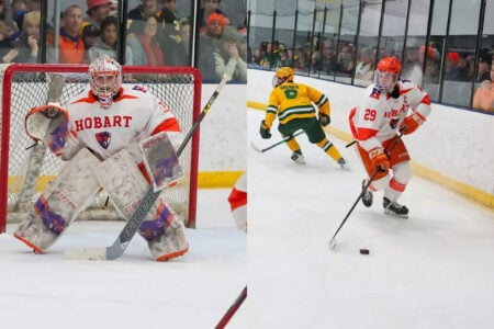Hobart hockey goalie positioned in net, and a Hobart player (29) skates with the puck past an opponent on an ice rink.