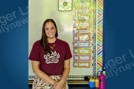 Young woman in a maroon "Pride Genesee" shirt smiles in a classroom with a pencil-themed daily schedule on a whiteboard.