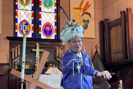 Indigenous person in feathered headdress and blue jacket, speaking from a church pulpit with stained glass.