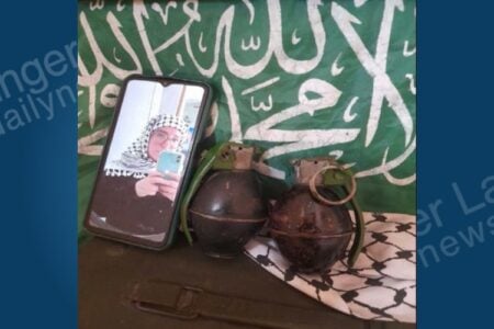Smartphone displays selfie of person in keffiyeh, next to two grenades, a keffiyeh cloth, and a green flag with Arabic script.