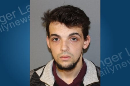 Young man with dark hair and beard, wearing a maroon shirt and black/grey patterned jacket, poses for a mugshot.