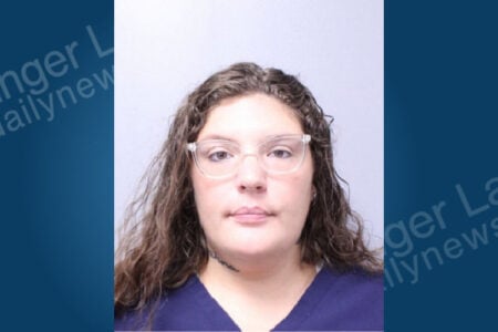 Woman with wavy brown hair, clear glasses, and dark blue top in a front-facing mugshot against a plain background.