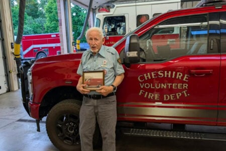 Firefighter holding a framed photo of a fire truck, standing by a Cheshire Volunteer Fire Dept. truck in a station.
