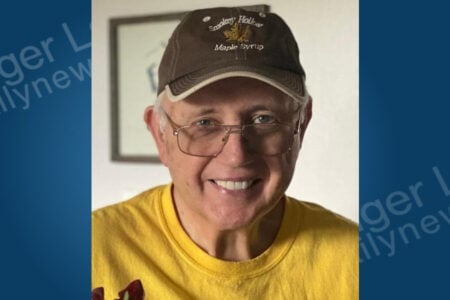 Smiling man in glasses and a brown "Smokey Hollow Maple Syrup" cap, wearing a yellow shirt, indoors.