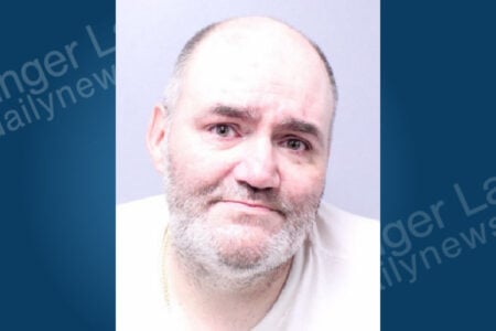 Bald man with a grey beard, white t-shirt, and gold chain looks forward against a light gray background.