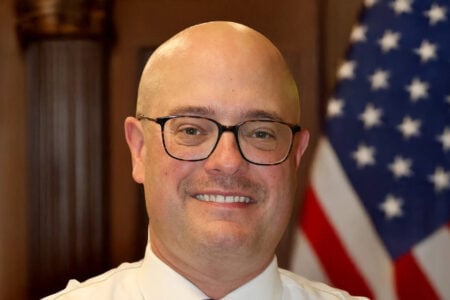Bald man in glasses and a white shirt smiling against a blurred American flag and column.