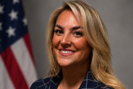 Headshot of a smiling blonde woman in a patterned navy blazer, with a blurry American flag behind her.