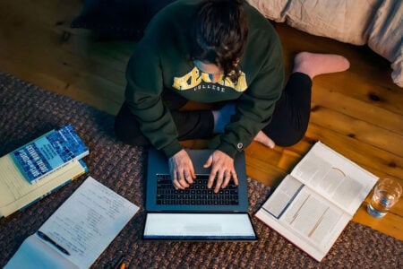 Keuka College student types on a laptop, surrounded by textbooks and notes, while sitting on a rug indoors.