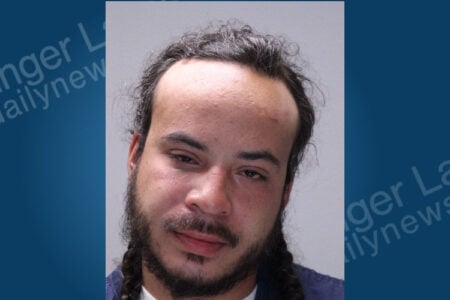 A man with dark curly hair, a beard, and two braids looks forward in a mugshot.