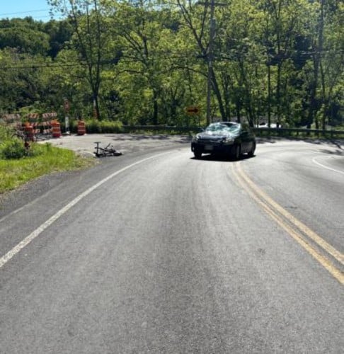 Dark car with shattered windshield and bicycle on ground after collision on a curved rural road.