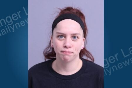 Mugshot of a woman with a black headband, reddish-brown hair in a messy bun, blue eyes, and pink ear plugs.