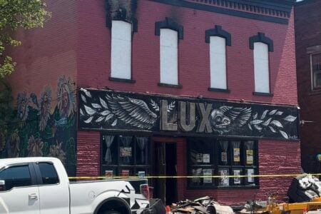 The red brick Lux building shows fire damage, charring above windows, debris on sidewalk, and a white truck.