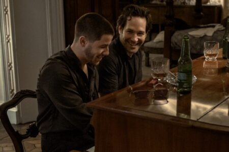 Nick Jonas and Paul Rudd share a laugh over drinks at a polished wooden table indoors.