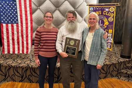 Walter Hurlbutt, holding an award plaque, smiles with two women in front of an American flag and Lions Club banner.
