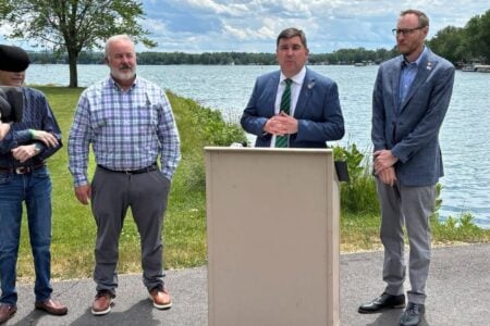 A man in a suit speaks at a podium outdoors by a lake, flanked by three other men including a videographer.