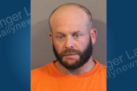 Bald man with a dark beard and blue eyes, wearing an orange shirt, looks directly at the camera in a mugshot.