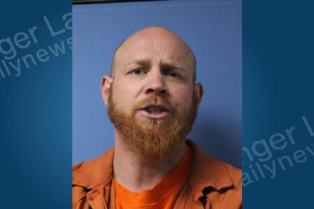 Mugshot of a bald man with a red beard and open mouth, wearing an orange shirt, against a plain blue background.