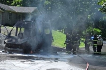 Firefighters douse a smoking, burnt van with foam on a suburban driveway next to a house.