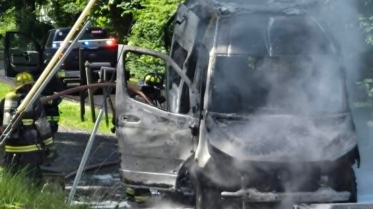 Firefighters hose down a severely burned van on a roadside, generating steam and smoke.