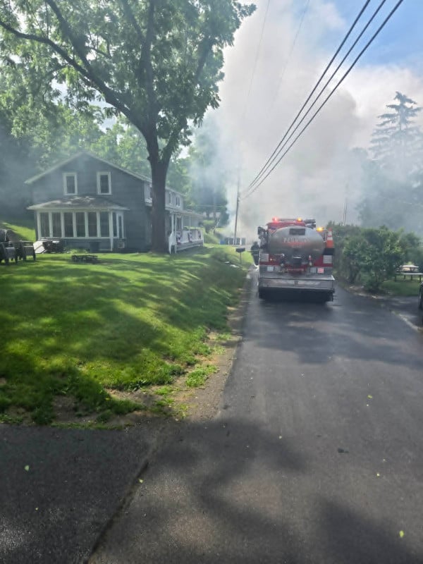 Phelps Fire tanker truck on a residential street with lights on, facing heavy smoke billowing from behind nearby homes.