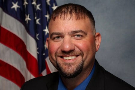 Smiling man with a beard, wearing a suit and blue shirt, against an American flag and a textured gray background.