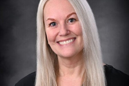 Headshot of a smiling woman with long blonde hair and blue eyes against a dark gray background.