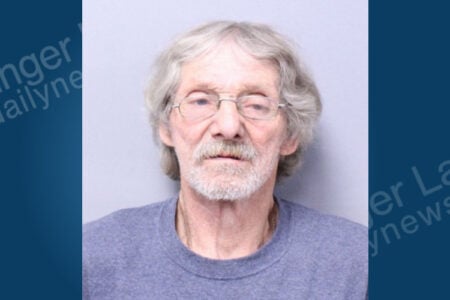 Older man with wavy gray hair, glasses, and a gray beard looks directly at the camera on a plain light background.