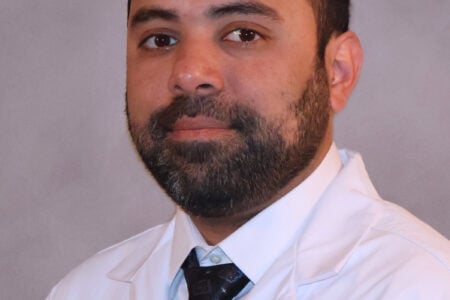 Bearded man in white UR MEDICINE Finger Lakes Health lab coat and tie, looking forward against a light background.