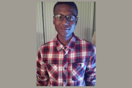 Young Black man in glasses and a red, white, and dark plaid shirt smiles against indoor blinds.