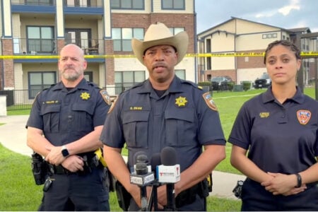 Sheriff E. Dean in a cowboy hat leads a press conference with two officers at an apartment complex crime scene.