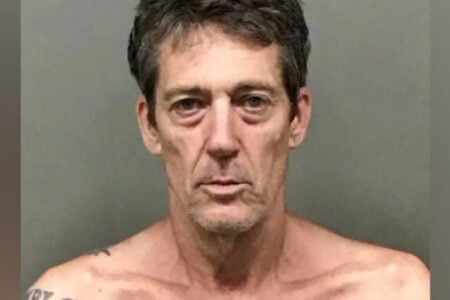 Mugshot of a man with disheveled brown hair, deep-set eyes, and a neutral expression, shirtless against a gray wall.
