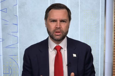 Professional man in navy suit, red tie, and American flag pin speaks animatedly in a modern studio.