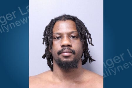 A Black man with dark dreadlocks and a beard looks straight ahead in a mugshot.