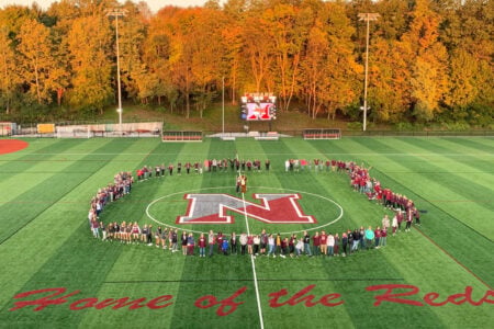 Students form a large 'N' on a green sports field with 'Home of the Reds' text and autumn trees.