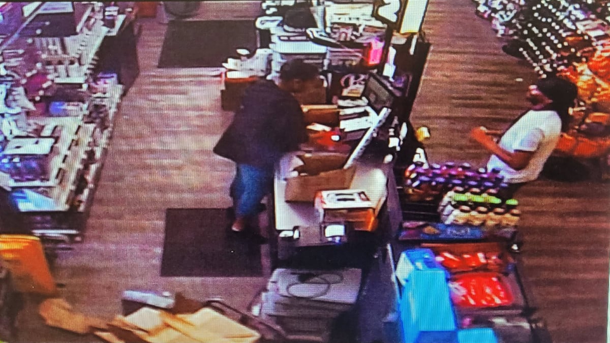 Surveillance shows two men in a convenience store: one rummages behind a counter, the other sits behind a display.