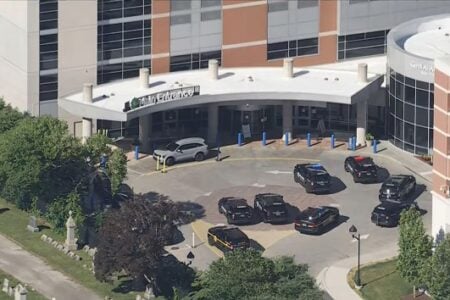 Multiple police cars parked outside the "Main Entrance" of Gene A. Adams hospital, with a cemetery visible on the left.