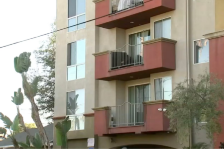 Multi-story apartment building with beige walls, reddish-brown balconies, and tall bird-of-paradise plants.