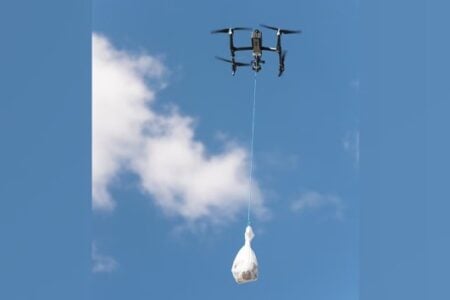 A black drone flies in a blue sky with white clouds, carrying a lumpy white bag suspended by a blue string.
