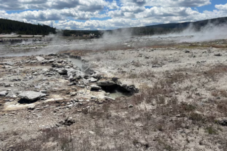 Yellowstone's geyser basin with steam rising from thermal features, a boardwalk and river in the background.
