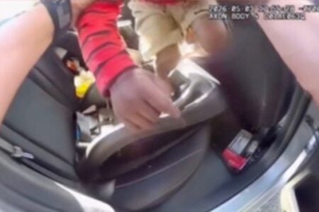 Body camera view shows an officer's arm reaching towards a child's legs in a car seat in the back of a vehicle.