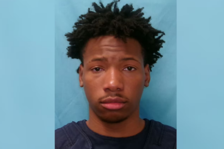 Young Black man with dark, curly hair, wearing a dark shirt, looks forward against a light blue background.