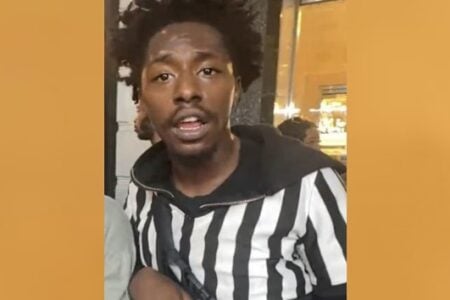 Black man in a black and white striped hoodie looks forward with mouth open, in a dimly lit setting.