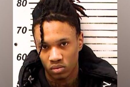 Young man with dreadlocks stares forward in a mugshot against a white height chart with black markings.