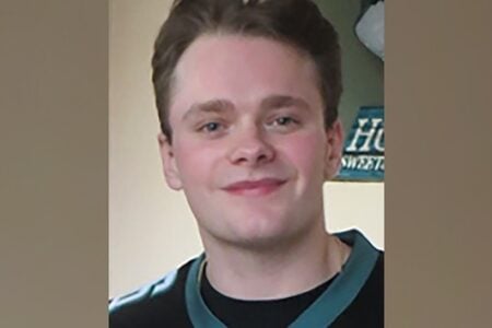 Young man with light hair and blue eyes smiles faintly, wearing a dark shirt with teal trim, against a plain background.