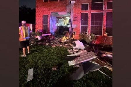 Damaged brick house at night with a large hole in its front wall, debris on the lawn, and a worker under emergency lights.