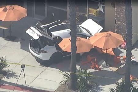 White SUV crashed into an outdoor dining area, overturning tables and orange umbrellas, with caution tape.